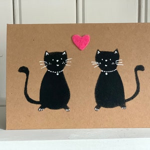 May include: A greeting card with a brown background features two black cat illustrations facing each other. Each cat has white whiskers and a white collar. A small pink heart is above the cats.