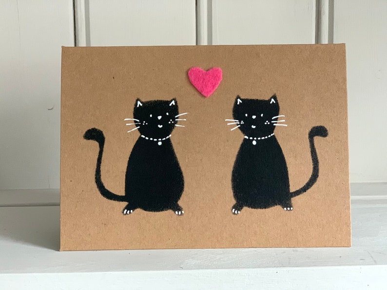 May include: A greeting card with a brown background features two black cat illustrations facing each other. Each cat has white whiskers and a white collar. A small pink heart is above the cats.