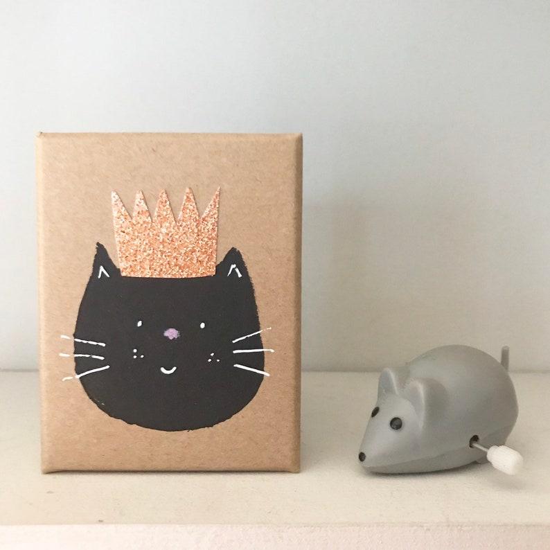 Puede incluir: Una caja de cart&oacute;n marr&oacute;n con un gato negro que lleva una corona de purpurina dorada. El gato tiene una nariz rosa y bigotes blancos. Un rat&oacute;n de juguete de cuerda gris est&aacute; en primer plano.