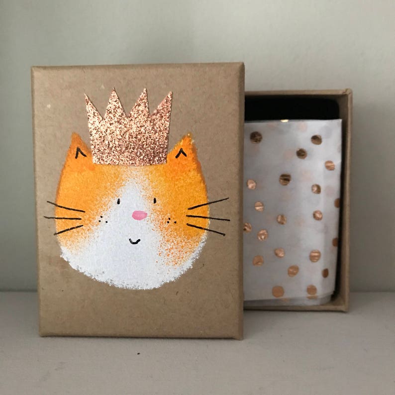 Puede incluir: Una peque&ntilde;a caja de cart&oacute;n con una ilustraci&oacute;n de un gato marr&oacute;n y blanco que lleva una corona dorada. La caja est&aacute; abierta y el interior est&aacute; forrado con papel de seda blanco con lunares dorados.