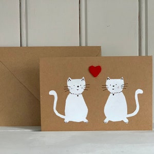 May include: A brown greeting card with two white cat illustrations and a red heart. The cats have black whiskers and are wearing black collars. An envelope is next to the card. The background is a white wooden surface.