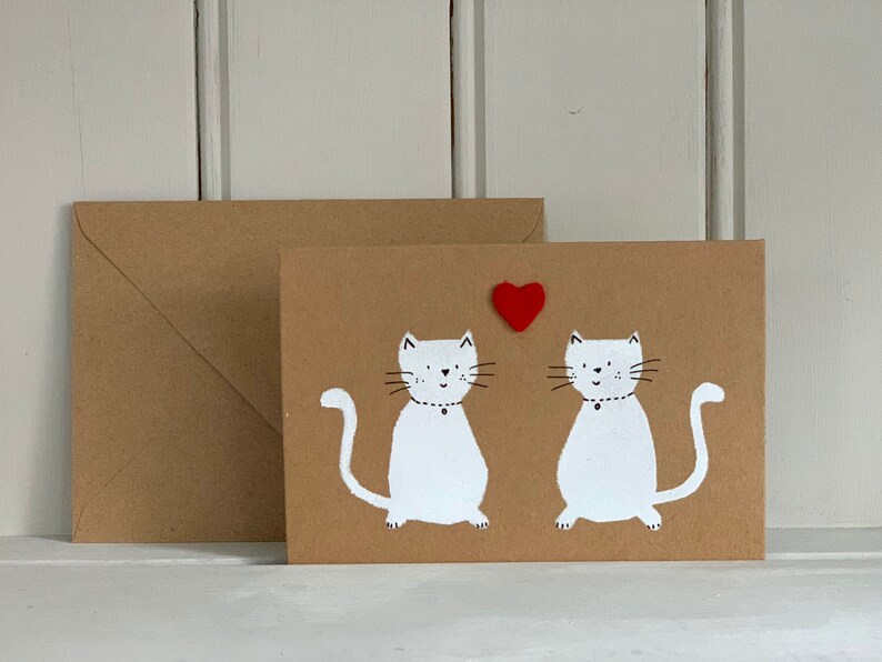 May include: A brown greeting card with two white cat illustrations and a red heart. The cats have black whiskers and are wearing black collars. An envelope is next to the card. The background is a white wooden surface.
