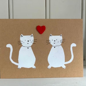 May include: A greeting card featuring two white cats with black whiskers and a red heart. The cats are on a brown background. Each cat has a black collar with a small black dot. The card is rectangular.