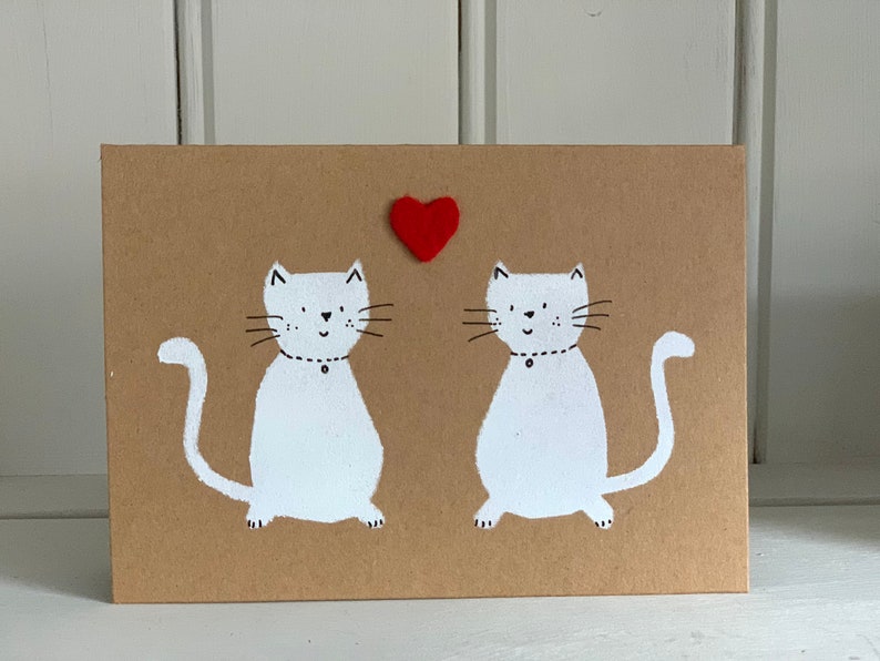 May include: A greeting card featuring two white cats with black whiskers and a red heart. The cats are on a brown background. Each cat has a black collar with a small black dot. The card is rectangular.