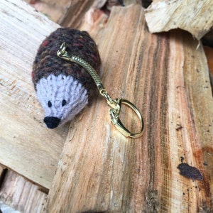 K&ouml;nnte beinhalten: Ein brauner und grauer gestrickter Igel-Schl&uuml;sselanh&auml;nger mit einem goldenen Metallring und einer Kette.