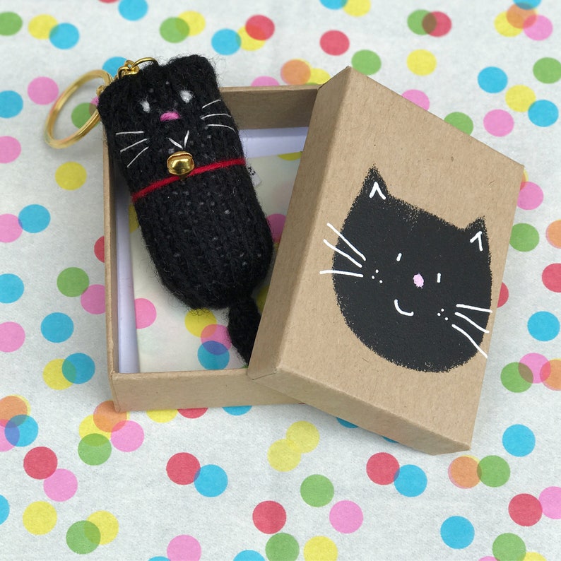Puede incluir: Un llavero de gato negro de punto con un collar rojo y una campana dorada, en una caja marr&oacute;n con una ilustraci&oacute;n de cara de gato negro.