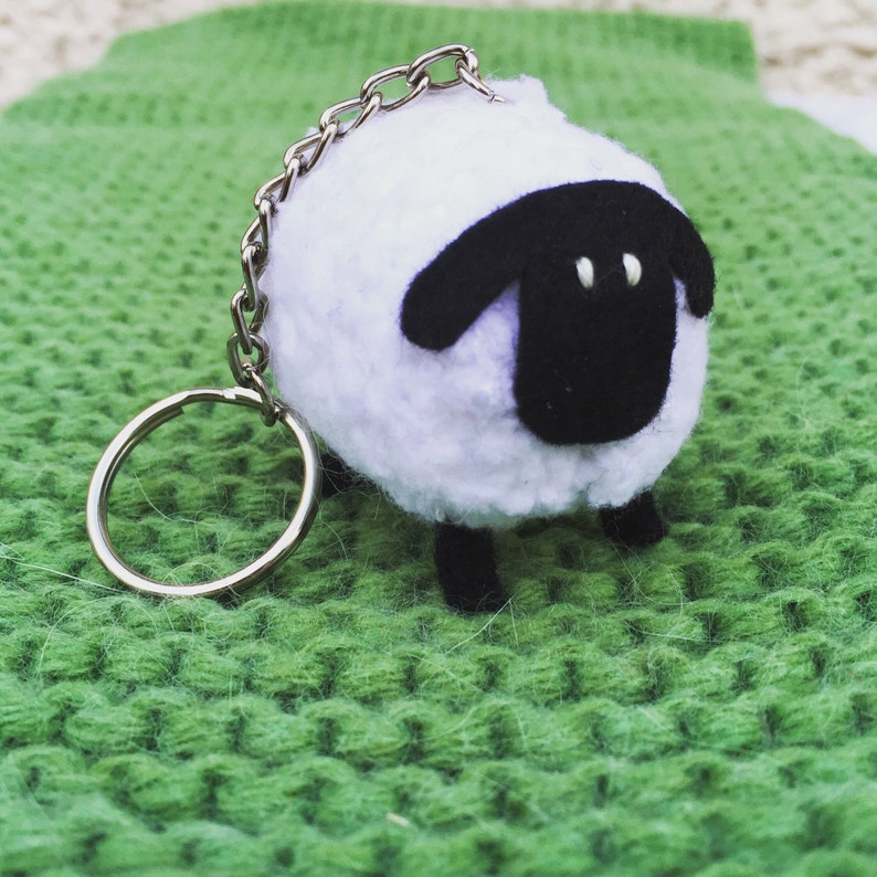May include: A white fluffy sheep keychain with black ears and a black face. The keychain has a silver metal ring and chain.