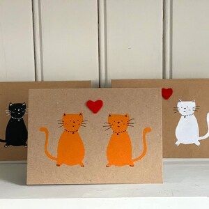 May include: Three handmade greeting cards with cat illustrations. One card features a black cat, another has two orange cats, and the third shows a white cat. Each cat has a simple design on a brown card.