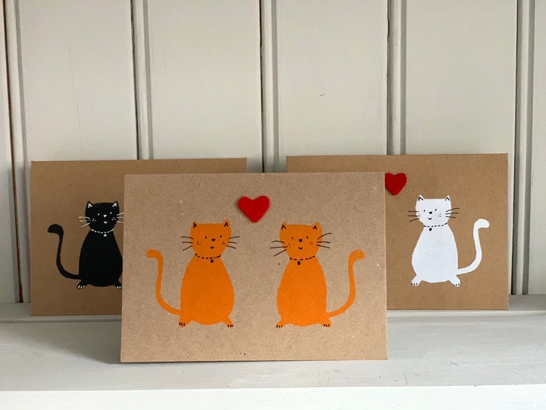 May include: Three handmade greeting cards with cat illustrations. One card features a black cat, another has two orange cats, and the third shows a white cat. Each cat has a simple design on a brown card.