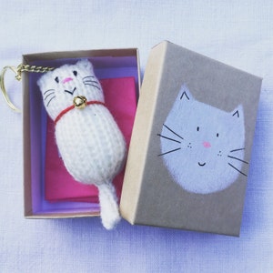 Puede incluir: Un llavero de gato blanco tejido con una cinta roja y una campanita dorada, dentro de una peque&ntilde;a caja marr&oacute;n con una cara de gato gris pintada a mano en la tapa.