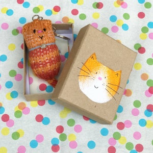 Puede incluir: Un peque&ntilde;o llavero de gato tejido de color naranja y rojo en una caja marr&oacute;n con una cara de gato naranja pintada a mano en la tapa.