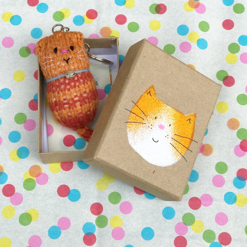 Puede incluir: Un peque&ntilde;o llavero de gato tejido de color naranja y rojo en una caja marr&oacute;n con una cara de gato naranja pintada a mano en la tapa.