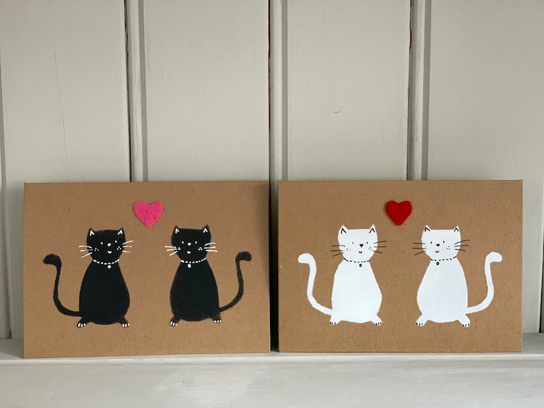 May include: Two cat-themed art pieces. One features two black cats with white details and a pink heart. The other shows two white cats with black details and a red heart. Both are on a brown background.