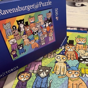 May include: A 500-piece Ravensburger puzzle featuring a colorful illustration of cartoon cats in various costumes. The puzzle box is dark blue with the Ravensburger logo and the word "Puzzle". The completed puzzle is partially assembled, showing more of the cat characters.