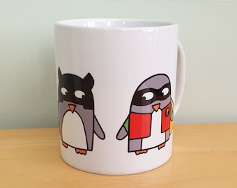 May include: White ceramic mug featuring two cartoon penguins. One penguin is dressed as a bat, the other as a superhero. The penguins are gray, black, red, and yellow. The mug is on a wooden surface.