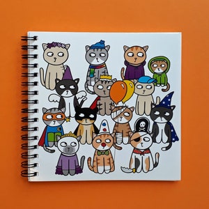 May include: Spiral-bound notebook featuring a whimsical illustration of cartoon cats in various costumes. The cats are dressed as a superhero, pirate, wizard, and more. The notebook is set against an orange background.