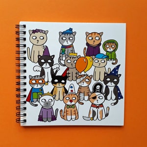 May include: A spiral-bound notebook featuring a colorful illustration of cartoon cats in various costumes. The cats are dressed as a superhero, a pirate, a wizard, and more. The notebook is on an orange background.