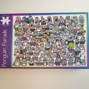 May include: A 500-piece jigsaw puzzle titled "Penguin Parade." The puzzle box features a colorful border and a purple background with the title in white. The puzzle image shows a large group of cartoon penguins in various outfits and poses.