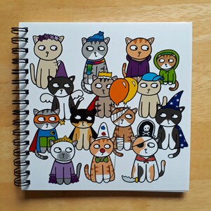 May include: A spiral-bound notebook featuring a collection of cartoon cats in various costumes. The cats are depicted in outfits such as a superhero, pirate, wizard, and princess. The notebook is white with a black spiral binding, set against a wooden background.