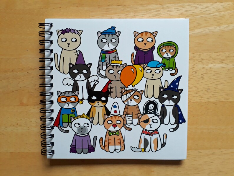 May include: A spiral-bound notebook featuring a collection of cartoon cats in various costumes. The cats are depicted in outfits such as a superhero, pirate, wizard, and princess. The notebook is white with a black spiral binding, set against a wooden background.