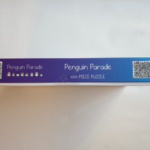 以下が含まれることがあります： 青と白のグラデーションデザインのパズルボックス。「Penguin Parade」と「1000 PIECE PUZZLE」の文字が表示されています。ボックスにはバーコードとパズル画像のコラージュが施されています。