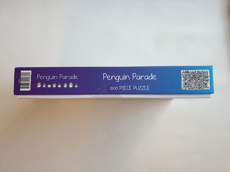 以下が含まれることがあります： 青と白のグラデーションデザインのパズルボックス。「Penguin Parade」と「1000 PIECE PUZZLE」の文字が表示されています。ボックスにはバーコードとパズル画像のコラージュが施されています。