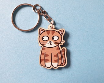 May include: A wooden cat keychain with a silver keyring and chain. The cat is brown with darker brown stripes and white paws. The cat is sitting and looking to the side. The background is a light blue.