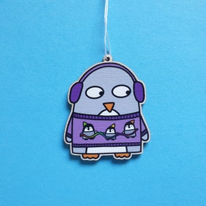 May include: Wooden penguin ornament with a gray body, purple earmuffs, and a purple sweater featuring three smaller penguins. The ornament has a white string for hanging and is set against a blue background.