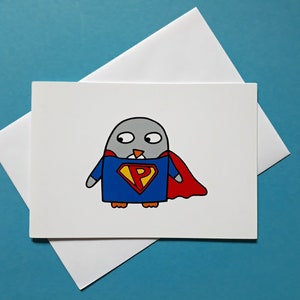 May include: A greeting card featuring a cartoon penguin dressed as a superhero. The penguin is gray with a blue and red costume, a red cape, and a yellow "P" on its chest. The card is white, with a white envelope.