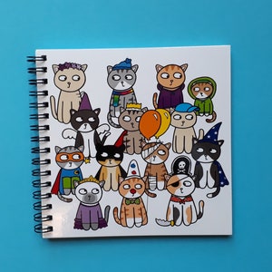 May include: A spiral-bound notebook featuring a colorful illustration of cartoon cats dressed in various costumes. The cats are depicted with different outfits, including a superhero, a pirate, and a wizard, on a white background.