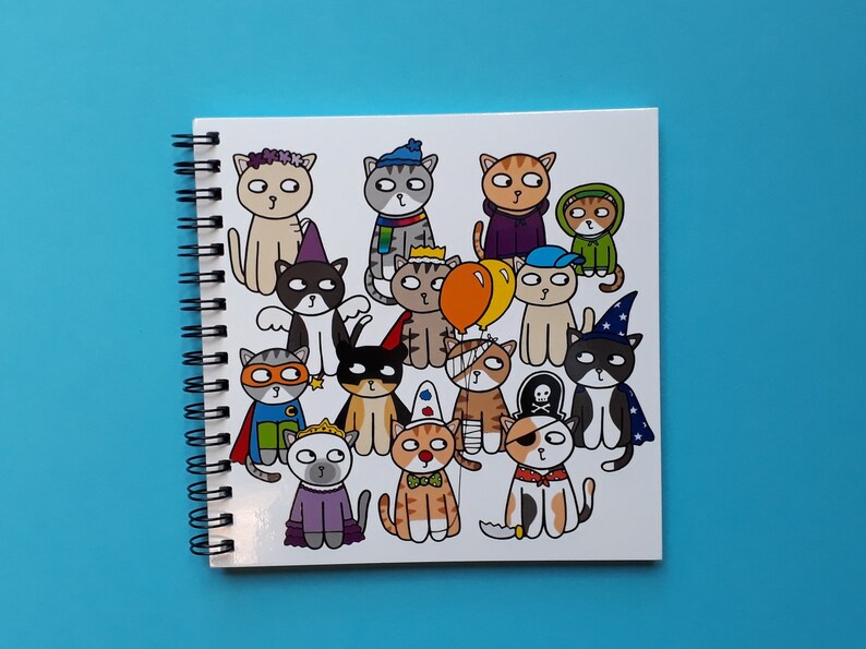 May include: A spiral-bound notebook featuring a colorful illustration of cartoon cats dressed in various costumes. The cats are depicted with different outfits, including a superhero, a pirate, and a wizard, on a white background.
