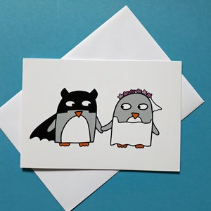 May include: A white greeting card with a cartoon illustration of two penguins holding hands. One is dressed as a superhero, the other in a bridal veil and flower crown. The card sits on a white envelope against a blue background.