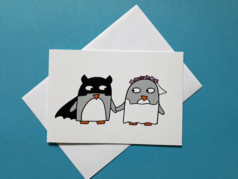 May include: A white greeting card with a cartoon illustration of two penguins holding hands. One is dressed as a superhero, the other in a bridal veil and flower crown. The card sits on a white envelope against a blue background.