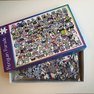 以下が含まれることがあります： 「Penguin Parade」というタイトルの500ピースのジグソーパズル。箱には、さまざまな衣装を着た多数の漫画のペンギンのカラフルなイラストが描かれています。パズルのピースは箱の中に表示され、組み立てる準備ができています。箱は長方形です。