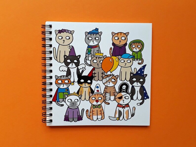 May include: A spiral-bound notebook featuring a colorful illustration of cartoon cats in various costumes. The cats are dressed as a superhero, a pirate, a wizard, and more. The notebook is on an orange background.