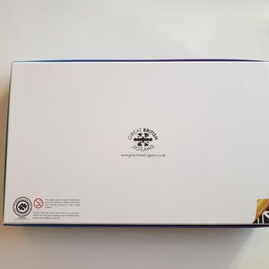 以下が含まれることがあります： 青と紫の縁取りが付いた白い長方形の箱。箱には「Great British Jigsaws」のロゴとウェブサイトのアドレスが表示されています。小さな部品に関する警告と、ユニオンジャックの旗の角も含まれています。