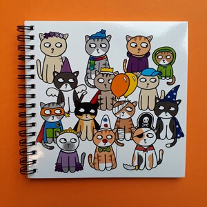 May include: A spiral-bound notebook with a white cover featuring a colorful illustration of cartoon cats in various costumes. The cats are dressed as a superhero, pirate, wizard, and more. The background is a solid orange.