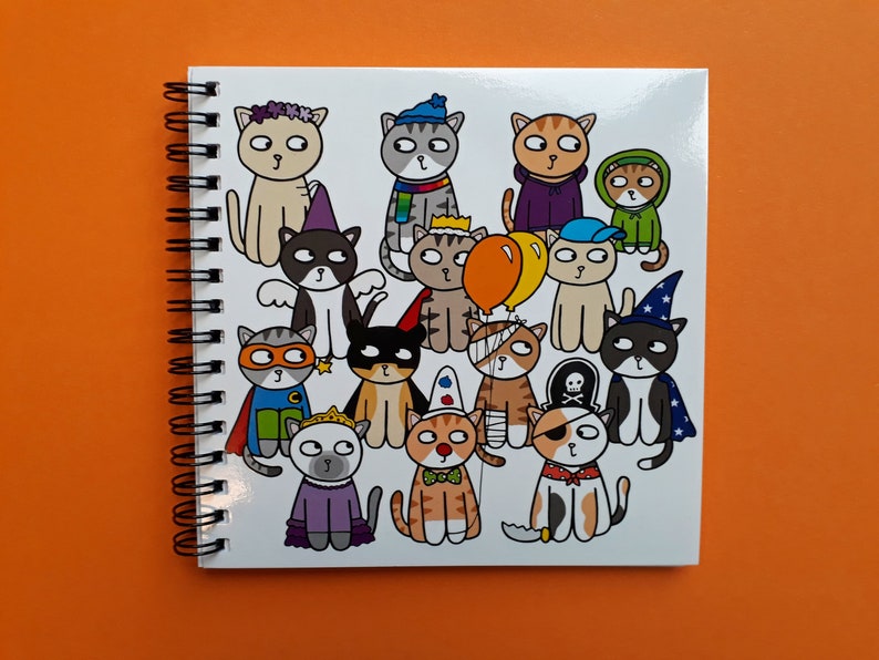 May include: A spiral-bound notebook with a white cover featuring a colorful illustration of cartoon cats in various costumes. The cats are dressed as a superhero, pirate, wizard, and more. The background is a solid orange.