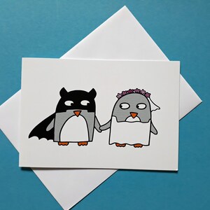 May include: A white greeting card with a cartoon illustration of two penguins. One is dressed as a superhero, the other as a bride. They are holding hands. The card is set against a blue background.
