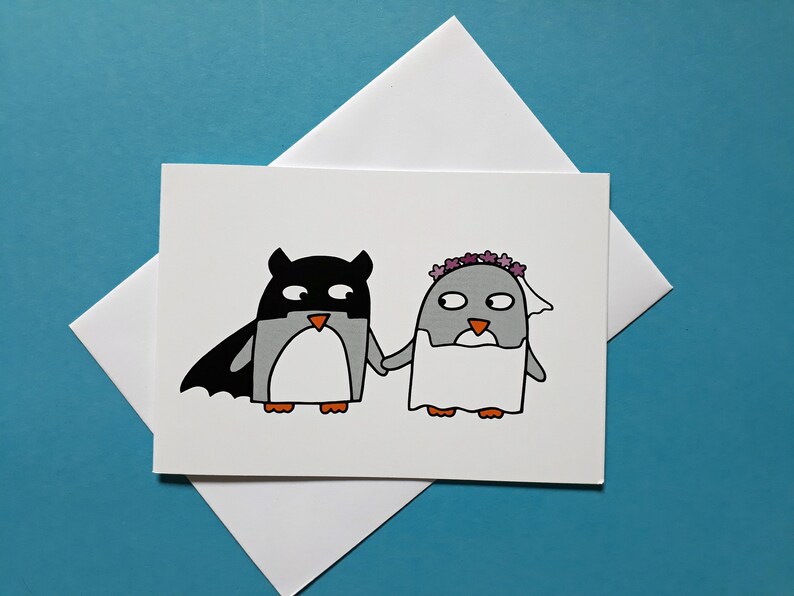 May include: A white greeting card with a cartoon illustration of two penguins. One is dressed as a superhero, the other as a bride. They are holding hands. The card is set against a blue background.
