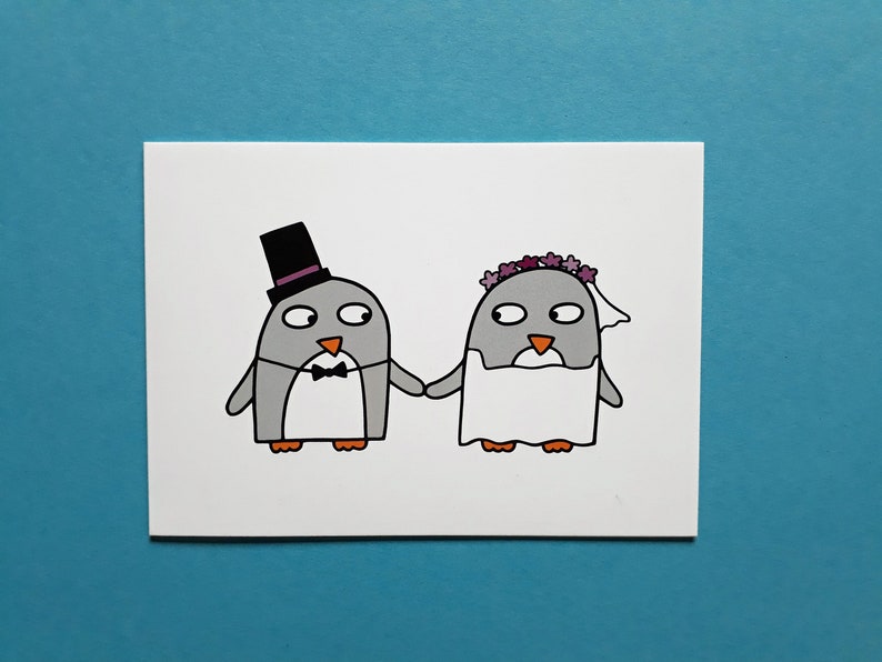 May include: A greeting card featuring a cartoon illustration of two penguins holding hands. One penguin wears a black top hat and bow tie, the other a floral crown and veil. The penguins are gray with orange beaks and feet.