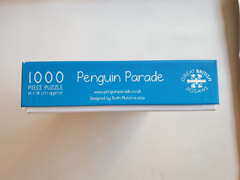 以下が含まれることがあります： 「1000 Piece Puzzle」と「Penguin Parade」の文字が入った青と白のパズルボックス。パズルのサイズは約69 x 48 cmです。ボックスには「Great British Jigsaws」の文字も含まれています。