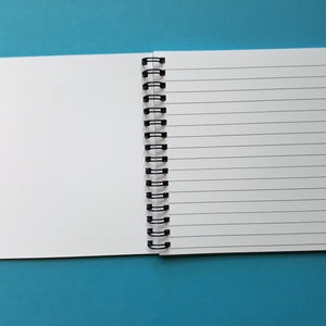 May include: Open spiral-bound notebook with blank white pages. One side is lined with horizontal black lines, the other is blank. The notebook is held together by a black metal spiral binding. The background is a solid teal color.