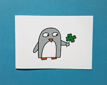 May include: A cartoon penguin illustration holding a green four-leaf clover. The penguin is grey with a white belly, orange feet and beak, and black outlines. The image is on a white background with a blue border.