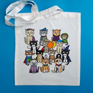 May include: White tote bag featuring a colorful illustration of cartoon cats dressed in various costumes. The cats are depicted with different outfits, including a superhero, a pirate, and a wizard. The bag has white handles and is set against a blue background.