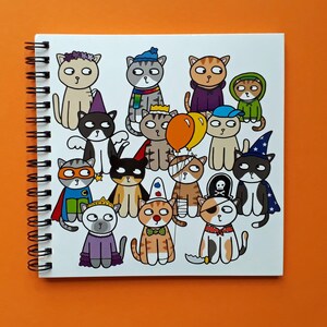 May include: A spiral-bound notebook featuring a collection of cartoon cats dressed in various costumes. The cats are depicted in a variety of outfits, including a superhero, a pirate, and a wizard. The notebook is on an orange surface.