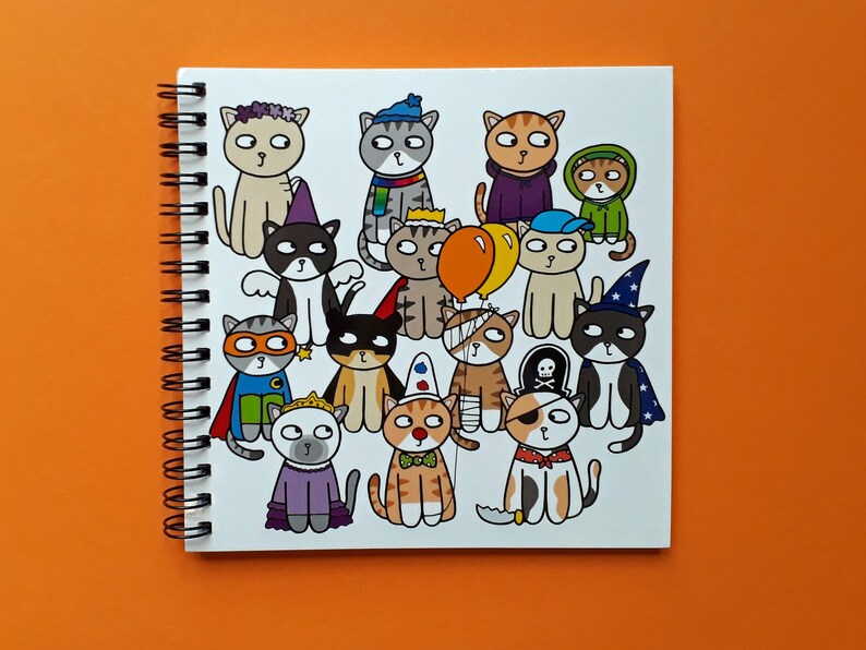 May include: A spiral-bound notebook featuring a collection of cartoon cats dressed in various costumes. The cats are depicted in a variety of outfits, including a superhero, a pirate, and a wizard. The notebook is on an orange surface.