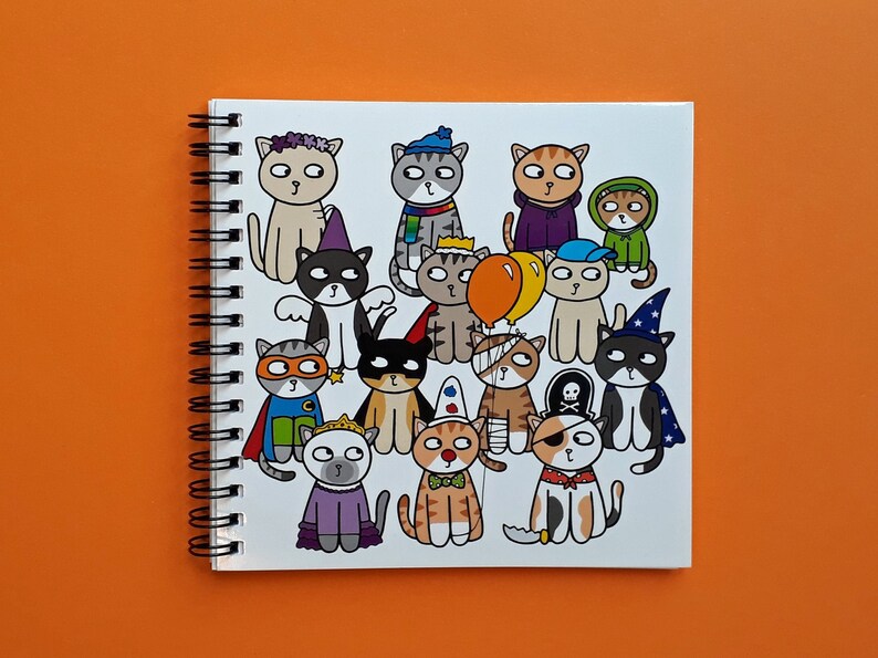 May include: Spiral-bound notebook featuring a whimsical illustration of cartoon cats in various costumes. The cats are dressed as a superhero, pirate, wizard, and more. The notebook is set against an orange background.