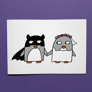 May include: A white card with a cartoon illustration of two penguins. One is dressed as a bat, the other as a bride. They are holding hands. The penguins are grey, black, and white with orange beaks and feet. The bride has a veil and floral crown.
