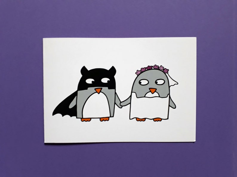 May include: A white card with a cartoon illustration of two penguins. One is dressed as a bat, the other as a bride. They are holding hands. The penguins are grey, black, and white with orange beaks and feet. The bride has a veil and floral crown.